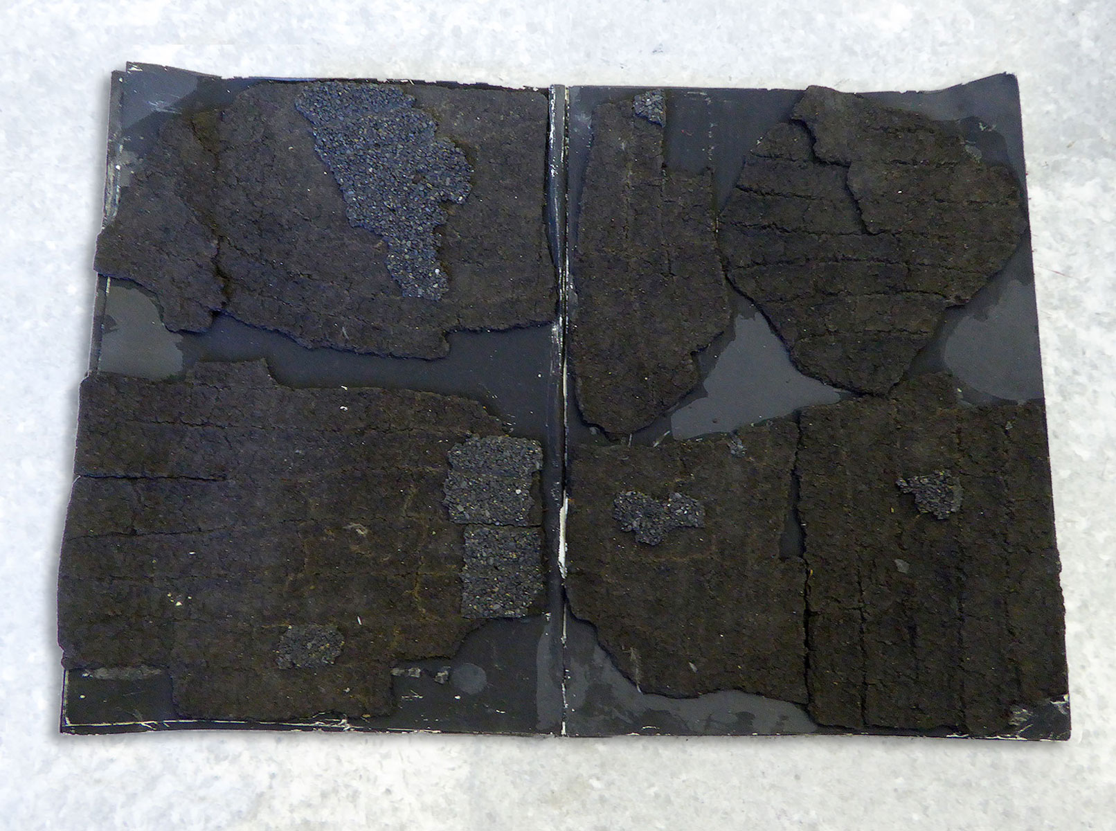 A dark, heavily textured mixed-media work resembling an open book, its two pages encrusted with tar-like black and deep blue material, photographed against pale grey concrete.